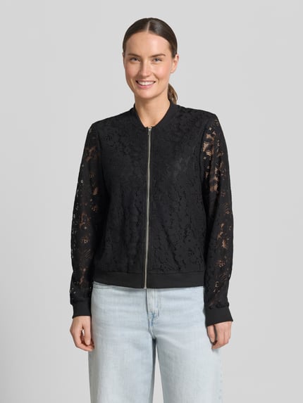 Kobiece model prezentuje Regular Fit Blouson aus Baumwoll-Mix Modell 'HONEY' od Vero Moda, czarny, wzór kwiatowy, materiał szydełkowy, kołnierz typu college, zamek błyskawiczny, prążkowane wykończenia, długi rękaw; ujęcie całej sylwetki z przodu.