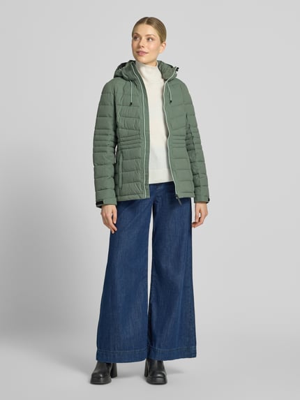 Frauenmodel trägt Jacke mit Kapuze und Label-Patch Modell von G.I.G.A. DX in Grün mit Reißverschluss, Ganzkörperansicht von vorne. Kontext: Damen Jacke Modell 'KOW10', gerader Schnitt, Kapuze mit Tunnelzug und Kordelstoppern, Reißverschluss, Label-Patch, seitliche Reißverschlusstaschen, Ärmelriegel mit Klettverschluss, 10.000 mm Wassersäule, Rückenlänge 66 cm, Schulterbreite 40 cm, Brustweite 47 cm, Ärmellänge 64 cm, Größe 36.