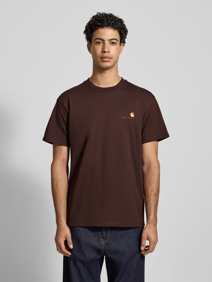 Männermodel trägt T-Shirt mit Label-Stitching von Carhartt Work In Progress in Braun, Reine Baumwolle, Regular-Fit, gerippter Rundhalsausschnitt, Ganzkörperansicht von vorne.