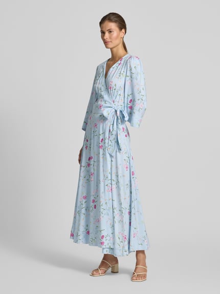Vrouwenmodel draagt lichtblauwe dames wikkeljurk met bloemenprint van YAS, zuiver lenzing™ ecovero™-vezels, getailleerd model, midi-jurk, V-hals, zijstrik, 3/4-mouw, ruglengte maat S 126 cm, borstwijdte 50 cm, mouwlengte 48 cm, model 'SAVANNA'. Ganzkörperansicht von voren.