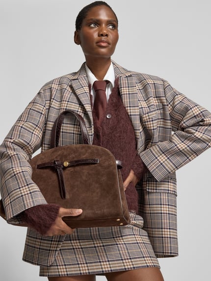 Frauenmodel trägt Crossbody Bag aus echtem Leder von Tory Burch in Dunkelbraun mit Karabinerverschluss und verstellbarem Riemen, kariertes Outfit, Ganzkörperansicht von vorne. Damen Crossbody Bag mit kurzem Henkel, Innenfach, 33x26x14 cm, strukturierte Oberfläche.