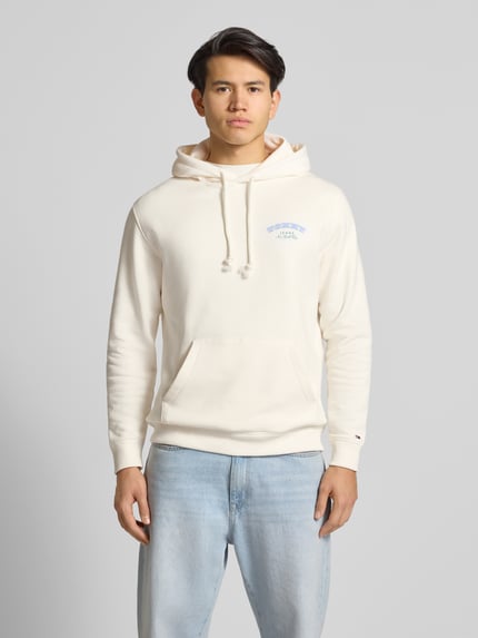 Męski model nosi Regular Fit Hoodie aus reiner Baumwolle od Tommy Jeans w kolorze jednolitym, z niebieskim napisem, kangurzą kieszenią, długimi rękawami i żebrowanymi wykończeniami. Minimalistyczna struktura bawełny typu sweat. Ganzkörperansicht von vorne.