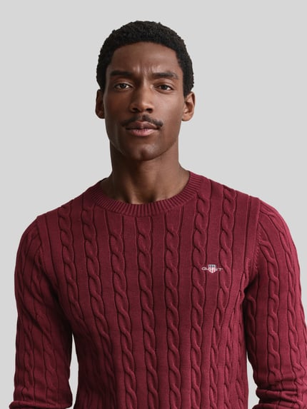 Männermodel trägt Regular Fit Pullover aus reiner Baumwolle von GANT in Dunkelrot mit Zopfmuster, Herren Pullover, Nahaufnahme Oberkörper.