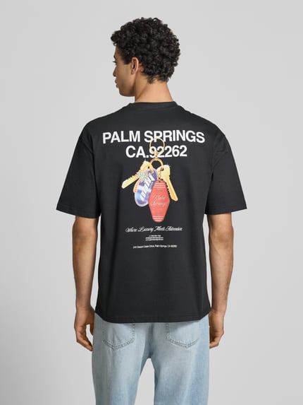 Männermodel trägt Boxy Fit T-Shirt aus reiner Baumwolle Modell 'FRED ESCAPE' von Only & Sons, schwarz, Motiv-Print, Ganzkörperansicht Rückansicht