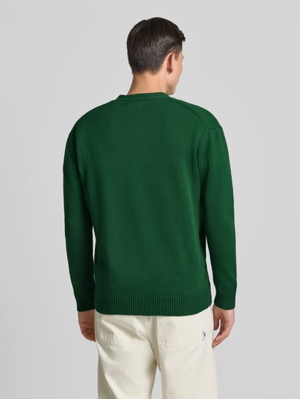 Männermodel trägt Strickjacke mit V-Ausschnitt Modell 'Tay' von Wood Wood aus grüner Baumwolle in geradem Schnitt, Ganzkörperansicht Rückansicht.