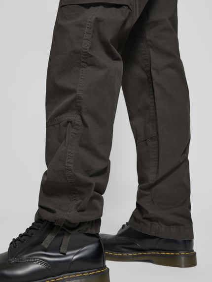 Männermodel trägt Straight Fit Cargohose mit Gesäßtaschen von G-Star Raw aus reiner Baumwolle, in dunklem Braun, Detailaufnahme Beine.