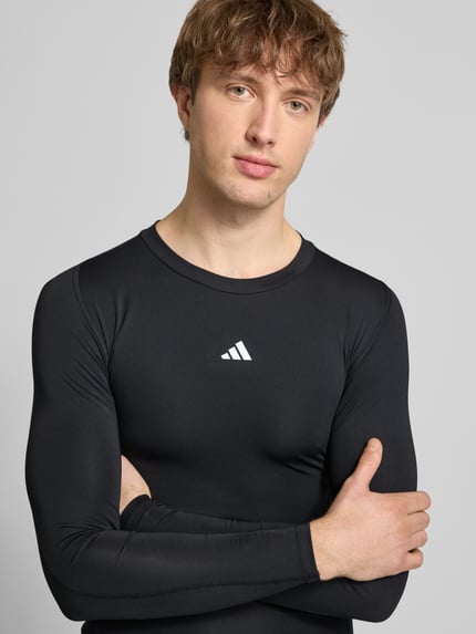 Męski model nosi Longsleeve mit Label-Print od adidas Training, czarny, elastyczny materiał, krój slim fit, z logo z przodu, zbliżenie na górną część ciała.