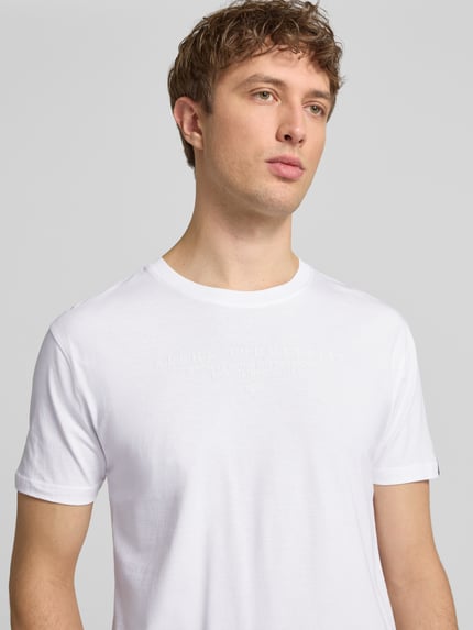 Männermodel trägt T-Shirt aus Baumwolle von Alpha Industries in Weiß mit Label-Details, Gerader-Schnitt, Rundhalsausschnitt, Nahaufnahme Oberkörper. Herren T-Shirt, Rückenlänge bei Größe M: 68 cm, Reine Baumwolle.