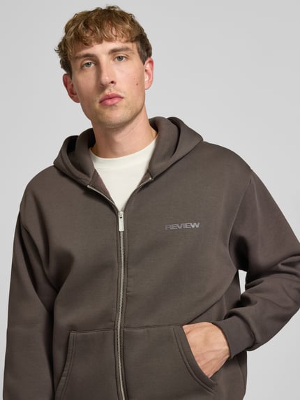 Männermodel trägt Relaxed Fit Sweatjacke aus Baumwoll-Mix mit Label-Detail von REVIEW in Braun, sichtbare Känguru-Tasche, Nahaufnahme Oberkörper