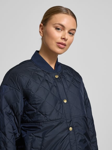 Frauenmodel trägt Relaxed Fit Steppjacke mit Rumpftunnelzug von Barbour in Optik: unifarben, Dunkelblau, kariertes Muster, Druckknopfverschluss, Langarm, Label-Schriftzug, College-Kragen, seitliche Eingrifftaschen, Innentasche, Label-Stitching, glatte strukturierte Oberfläche, Nahaufnahme Oberkörper.