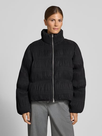 Frauenmodel trägt schwarze Steppjacke mit Zweiwege Reißverschluss von YAS, unifarbene Optik, Stehkragen, Strukturmuster, Ganzkörperansicht von vorne.