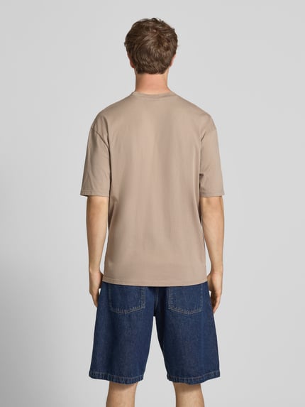 Männermodel trägt Oversized T-Shirt mit Label-Stitching von Pegador in Beige, kombiniert mit dunkelblauer Jeansshorts, Ganzkörperansicht Rückansicht. Oversized Herren T-Shirt aus Baumwoll-Mix mit Rundhalsausschnitt, Rückenlänge Größe M 70 cm.