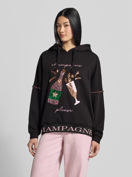 Frauenmodel trägt Hoodie aus reiner Baumwolle von miss goodlife in Schwarz mit Pailletten-Besatz, lockere Passform, elastische Abschlüsse, Ganzkörperansicht von vorne.