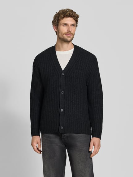 Męski model nosi kardigan aus Kaschmir-Mix mit V-Ausschnitt von FTC Cashmere, mieszanka kaszmiru z dodatkiem streczu, prosty krój, dekolt w serek, listwa guzikowa, Długość tyłu w rozmiarze 50: 73 cm, Długość rękawów w rozmiarze 50: 62 cm, kolor czarny, faktura prążkowana. Ganzkörperansicht von vorne.