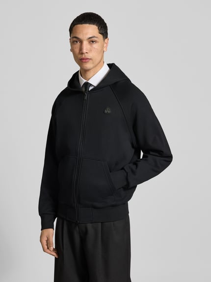 Männermodel trägt Sweatjacke mit Kapuze von Moose Knuckles, schwarzer Baumwoll-Mix, lockerer Schnitt, Kängurutasche, Ganzkörperansicht von vorne.