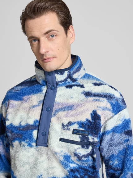 Męski model nosi Regular Fit Pullover mit Logo-Patch od Columbia, efekt fließ, naszywka z logo, brystasza z zamkiem, stójka z listwą z zatrzaskami, elastyczne wykończenia, niebiesko-biały wzór, zbliżenie na górną część ciała