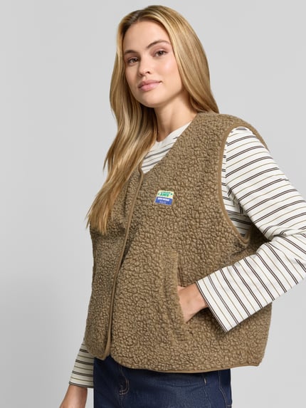 Frauenmodel trägt Regular Fit Weste aus Teddyfell Modell 'Hoktown' von American Vintage, beige, strukturierte Oberfläche, Nahaufnahme Oberkörper. Damen Weste aus Teddyfell, Logo-Patch und Knopfleiste im Fokus, Kontext: Rückenlänge 57 cm, Schulterbreite 43 cm, Brustweite 63 cm.