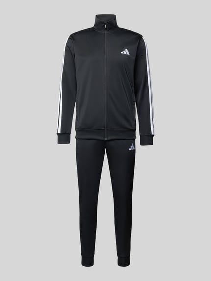 Männermodel trägt Trainingsanzug mit Stretch-Anteil von adidas Sportswear in Schwarz mit weißen Label-Details, Elastischer-Material-Mix, Gerade Passform, Stehkragen, Durchgehender Reißverschluss, elastische Abschlüsse, Elastikbund, Eingrifftaschen, Ganzkörperansicht von vorne.