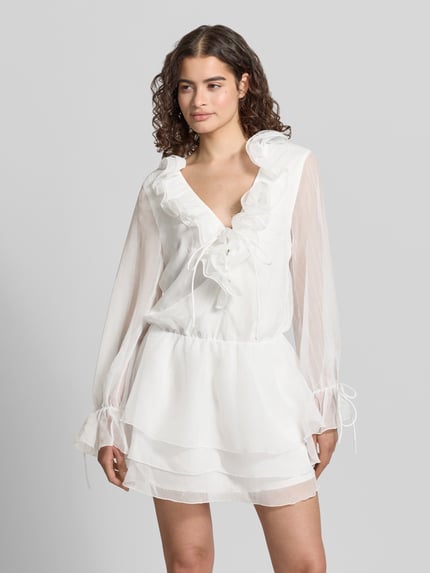 Vrouwenmodel draagt Minikleid met Rüschen und V-Ausschnitt van Gina Tricot in wit, getailleerd model, mouwen met vetersluiting, ganzkörperansicht van voren, ruglengte maat XS 85 cm, borstwijdte maat XS 49 cm, mouwlengte maat XS 64 cm