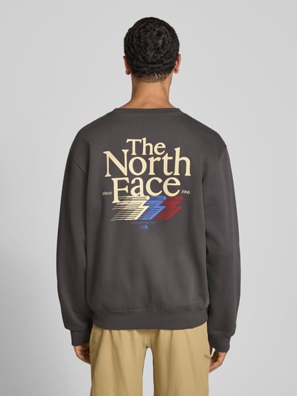 Männermodel draagt Relaxed Fit Sweatshirt aus katoenmix van The North Face met logoprint en ronde hals, herensweatshirt, ruglengte maat M 68 cm, mouwlengte maat M 68 cm, elastische afwerking, donkergrijs met meerkleurig logo, ganzlichaamsaanzicht van achteren