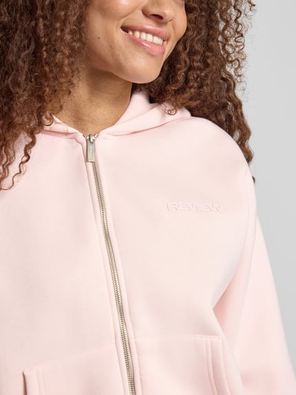 Vrouwenmodel draagt Relaxed Fit Sweatjacke mit Kapuze van REVIEW, zachte roze kleur, katoen, logo-print, kangoeroezakken, detailopname bovenlichaam.