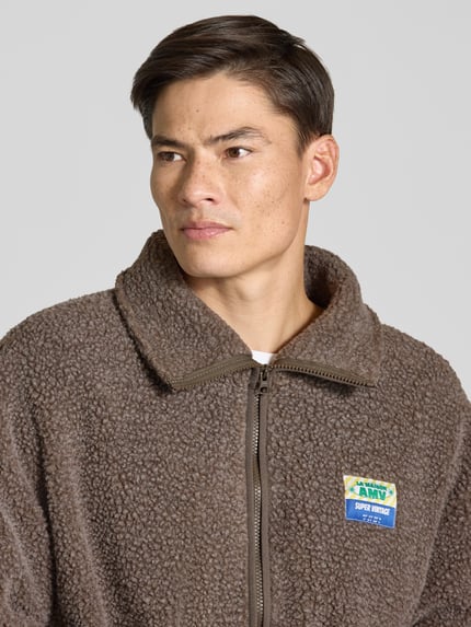 Męski model nosi Fleecejacke mit Label-Detail Modell 'HOKTOWN' od American Vintage, kolor brązowy, tekstura teddy, krój regular fit, stójka, zamek błyskawiczny, detal z logo, szerokość w rozmiarze M/L: 67 cm, długość tyłu: 72 cm, szerokość ramion: 58 cm, długość rękawa: 55 cm, zbliżenie na górną część ciała.
