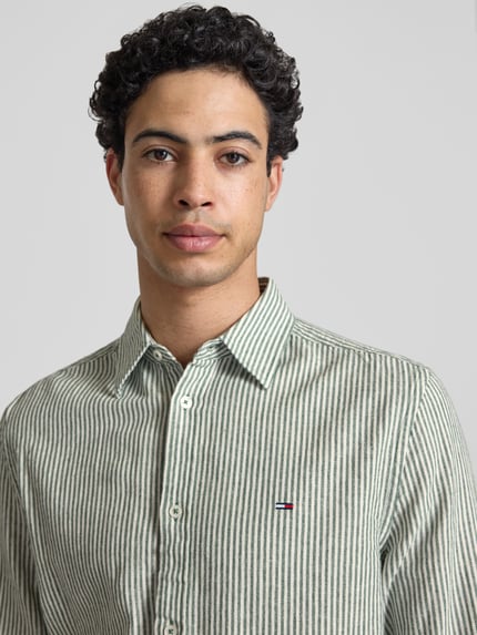 Männermodel trägt Regular Fit Freizeithemd aus Leinen-Baumwoll-Mix von Tommy Jeans, grün-weiß gestreift, Logo, Nahaufnahme Oberkörper.