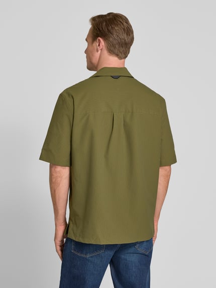 Męski model nosi Relaxed Fit Freizeithemd aus Seersucker-Material od Tommy Hilfiger w oliwkowej zieleni z drobnym wzorem, miękka tekstura, tylny karczek z kontrafałdą, krótkie rękawy, luźny krój, widok całej sylwetki z tyłu