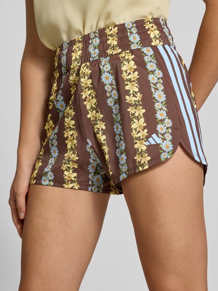 Frauenmodel trägt Regular Fit Sportshorts mit Allover-Muster Modell 'FARM SHORTS' von adidas, braun mit gelbem und blauem Muster, Damen Sportshorts von adidas Sportswear, Nahaufnahme Oberkörper.