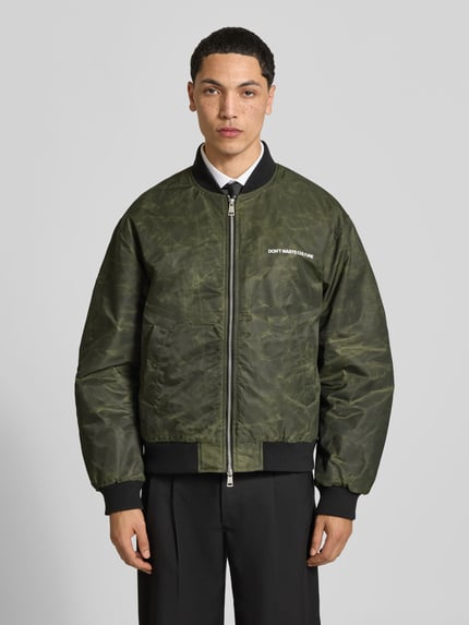 Männermodel trägt bomberka 'Bomberjacke mit Label-Print' von Don´t Waste Culture w kolorze khaki z nadrukiem z logo, krój regular fit, zamek błyskawiczny dwustronny, elastyczne wykończenia, stójka, długość tyłu w rozmiarze M: 67 cm, szerokość ramion w rozmiarze M: 53 cm, długość rękawów w rozmiarze M: 64 cm, Ganzkörperansicht von vorne
