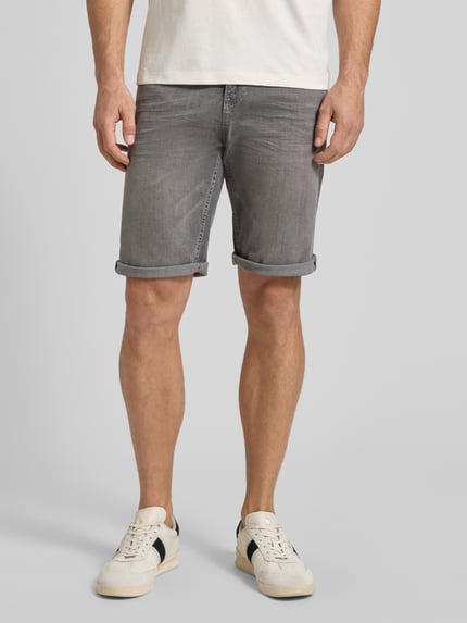 Mężczyzna nosi Regular Fit Jeans Bermudas aus Baumwoll-Mix od Tom Tailor, stylizowana na denim, krój regular fit, mieszanka bawełny, 5 kieszeni, aplikacja z logo, szlufki na pasek, zapięcie na guzik i ukryty zamek błyskawiczny, widok całej sylwetki z przodu.