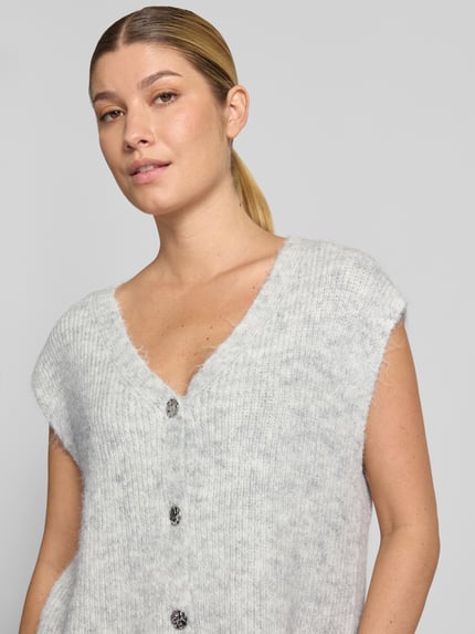 Kobiece model nosi kamizelkę dzianinową Strickweste mit V-Ausschnitt Modell 'Xandra' von Neo Noir, mieszanka materiałów z bawełną, krój regular fit, dekolt w serek, listwa guzikowa, długość tyłu w rozmiarze 36: 58 cm, zbliżenie na górną część ciała.