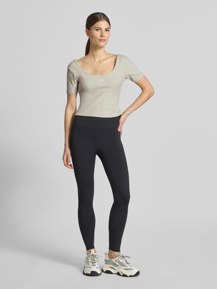 Frauenmodel trägt Cropped T-Shirt aus Lyocell-Mix Modell 'Circe' von Guess Activewear in Beige, kurzer Schnitt, Logo-Stitching, Ganzkörperansicht von vorne.
