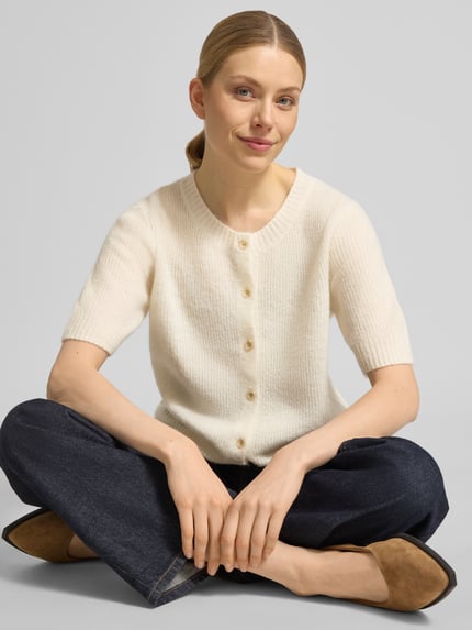 Modelka kobieca ma na sobie Cardigan aus Woll-Mix mit 1/2-Arm Modell 'Thora' od MOS MOSH, kolor kremowy, krój regular fit, okrągły dekolt, mieszanka wełny z dodatkiem alpaki, długość tyłu w rozmiarze S: 59 cm, długość rękawa: 30 cm, tekstura dzianiny, proste ciemnoniebieskie spodnie, całe ciało widoczne od przodu.