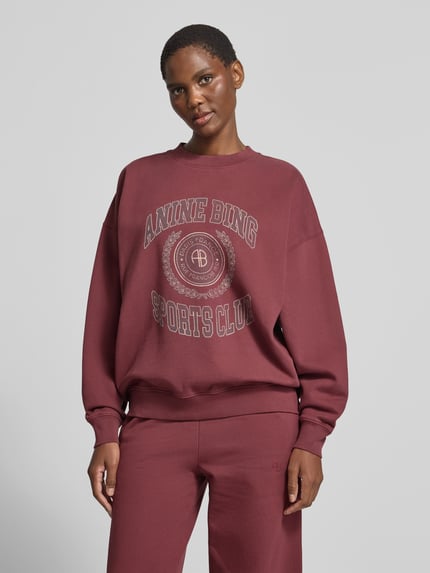 Frauenmodel trägt bordeauxrotes Sweatshirt mit Logo-Print Modell 'MILES SWEATSHIRT' von Anine Bing, Motiv-Print, Rundhals, unifarben, langarm, gerippte Abschlüsse, Ganzkörperansicht von vorne.