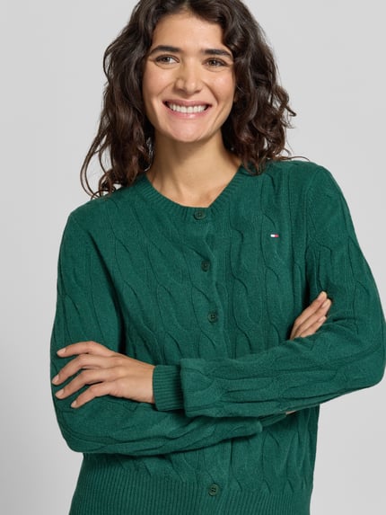Frauenmodel trägt Strickjacke aus Wolle mit Logo-Detail von Tommy Hilfiger in Grün mit Zopfmuster, Rundhals, Knopfleiste, Nahaufnahme Oberkörper.