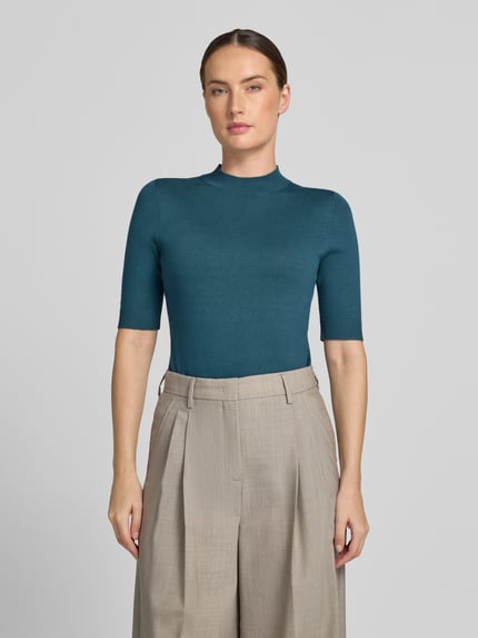 Frauenmodel trägt Slim Fit Strickpullover aus Viskose-Mix von COMMA in Blaugrün, feine Struktur, gerippte Abschlüsse, Halbarm, Ganzkörperansicht von vorne.