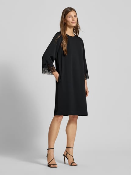Kobiece modelka nosi sukienkę Knielanges Kleid mit Rundhalsausschnitt Modell 'Dancia' von JOOP! Collection, czarny kolor, krój relaxed fit, okrągły dekolt, długość do kolan, rękaw o dł. 3/4 z obszyciem koronką, długość tyłu 100 cm w rozmiarze 36, szerokość klatki piersiowej 49 cm, długość rękawa 65 cm, gładka tekstura, całkowity widok z przodu.