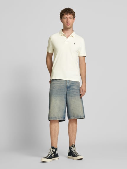 Męski model nosi Modern Fit Poloshirt mit Logo-Stitching od REVIEW, bawełniany miks, polokrag, krótka listwa guzikowa, długość pleców w rozmiarze M: 65 cm, jasnokremowy, jeanowe szorty, granatowe trampki, pełny widok z przodu.