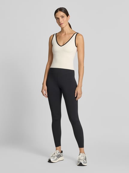 Frauenmodel trägt Slim Fit Top mit V-Ausschnitt von aim'n, cremefarbene Front, schwarze Konturen, elastischer Material-Mix, Ganzkörperansicht von vorne, Damen Top von aim'n, Rückenlänge bei Größe S 26 cm, Logo-Detail.