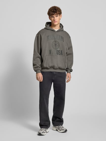 Männermodel trägt Hoodie mit Label-Print und Känguru-Tasche von KARL KANI, grauer Baumwoll-Mix, oversized, Ganzkörperansicht von vorne.