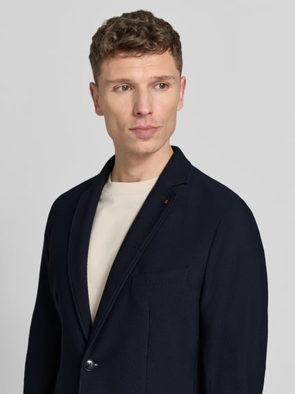 Männermodel trägt marynarkę na 2 guziki z mieszanki materiałów z dodatkiem streczu o kroju slim fit, fakturowany wzór, kołnierz z połami, nakładane kieszenie, długość tyłu w rozmiarze 50: 73 cm, szerokość ramion w rozmiarze 50: 42 cm, długość rękawów w rozmiarze 50: 47 cm, model 'Bob' od Zuitable; ciemnogranatowa, prosta faktura, gładka powierzchnia. Nahaufnahme górnej części ciała.