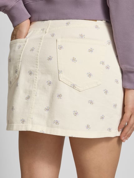 Frauenmodel trägt Ausgestellter Mini-Rock aus reiner Baumwolle Modell 'NAOMI' von Only mit Florales-Allover-Muster, Twill, Label-Patch, Knopfleiste, Gesäßtaschen und seitliche Eingrifftaschen, Creme mit kleinen Blumen, Nahaufnahme Rückansicht.