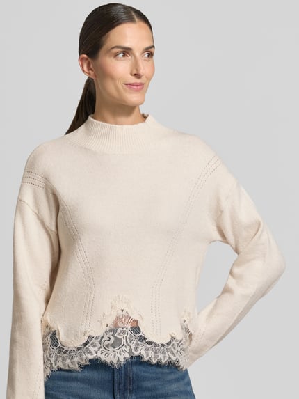 Frauenmodel trägt Regular Fit Strickpullover aus reiner Baumwolle mit Spitzensaum von GUESS, cremefarben, dezente Struktur, Nahaufnahme Oberkörper.