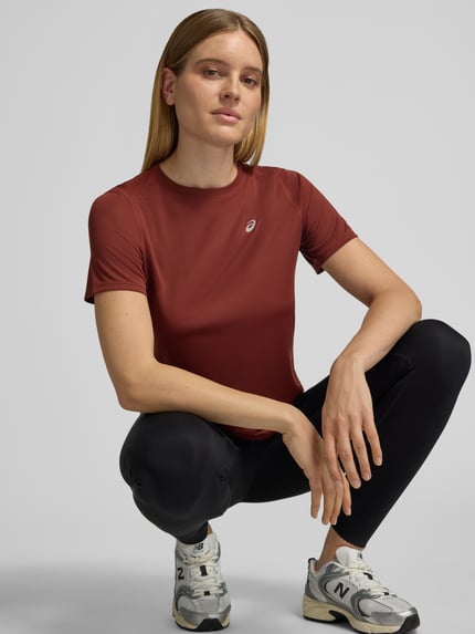 Modelka kobieca nosi Regular Fit T-Shirt mit Logo-Print od Asics w kolorze bordowym, okrągły dekolt, krótkie rękawy, długość tyłu w rozmiarze S: 63 cm, prosta struktura materiału, widok całej sylwetki z przodu.