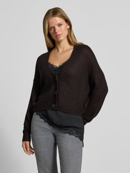 Frauenmodel trägt Boxy Fit Strickjacke mit V-Ausschnitt Modell 'CAROL' von ONLY, dunkles Strickmuster, kombiniert mit schwarzem Top und grauer Hose, Damen Strickjacke von ONLY, Ganzkörperansicht von vorne.