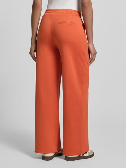 Frauenmodel trägt Wide Fit Stoffhose mit Viskose-Anteil Modell 'Kate' von ICHI in Orange, elastischer Material-Mix, Bundfalten, Ganzkörperansicht Rückansicht.