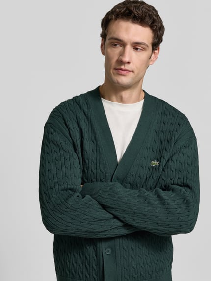 Männermodel trägt Cardigan mit Logo-Detail von Lacoste in grüner Strickoptik mit Zopfmuster, Nahaufnahme Oberkörper.