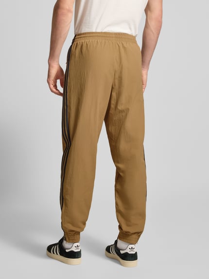 Männermodel trägt Sweatpants mit elastischem Bund von adidas Originals in Camel mit Reißverschlusstaschen und Label-Stitching, Lockerer-Schnitt, Ganzkörperansicht Rückansicht. Herren, Innenbeinlänge bei Größe M: 77 cm.