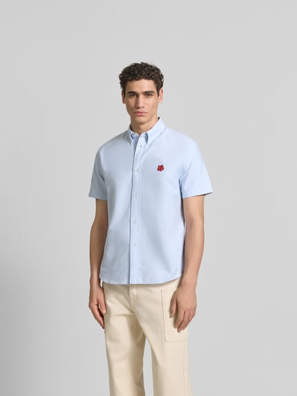 Męski model ma na sobie Slim Fit Freizeithemd mit Logo-Stitching od Kenzo z bawełny, krój slim fit, kołnierzyk typu button down, wyhaftowane logo, krótki rękaw, jasnoniebieski kolor, długość tyłu w rozmiarze M 49 cm, szerokość ramion w rozmiarze M 77 cm, długość rękawa w rozmiarze M 29 cm, gładka struktura, widok całej sylwetki z przodu.